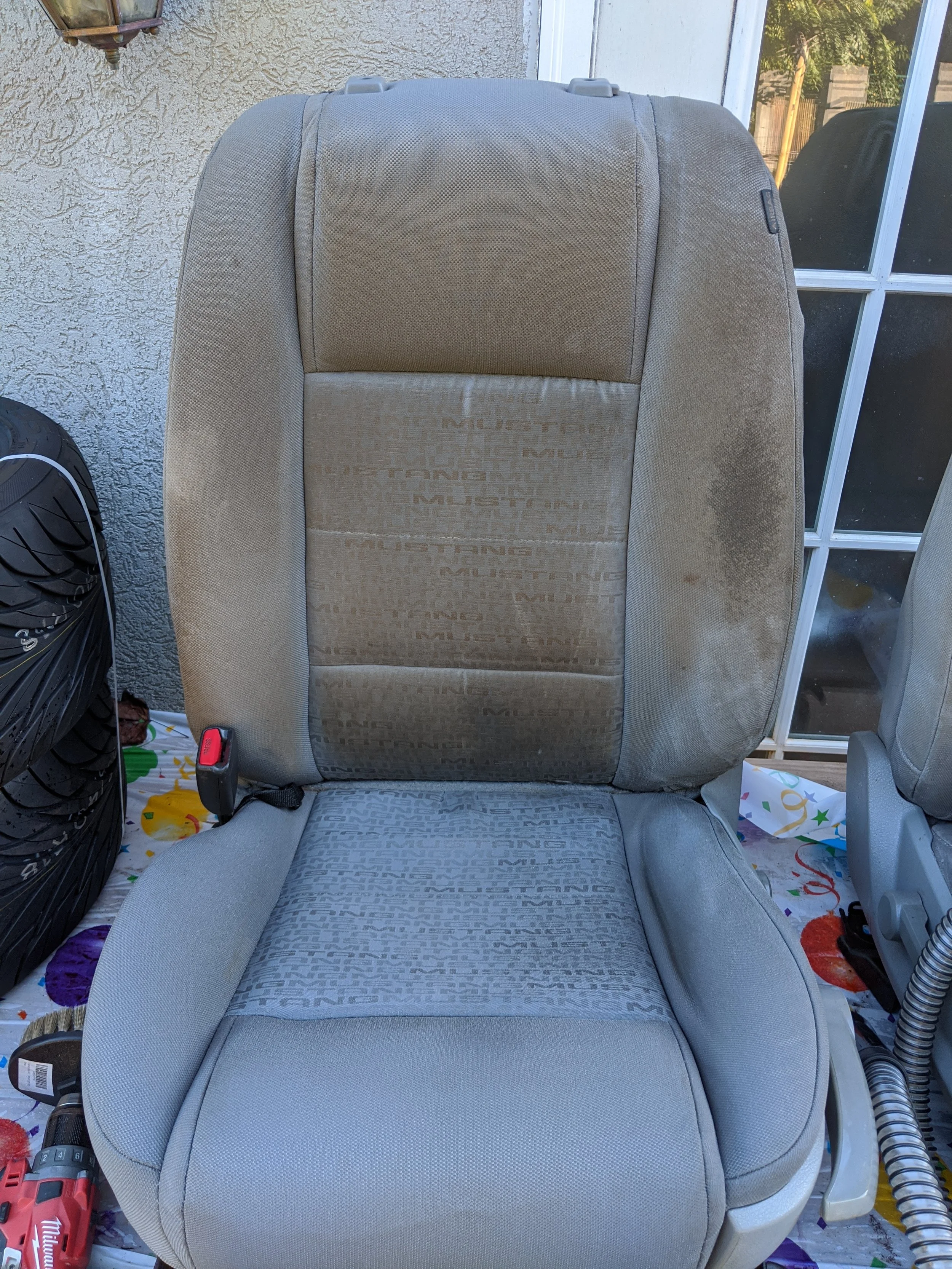 Car seat with mustard stains on the right shoulder area and some dirt marks, situated outdoors near a window and a tire.