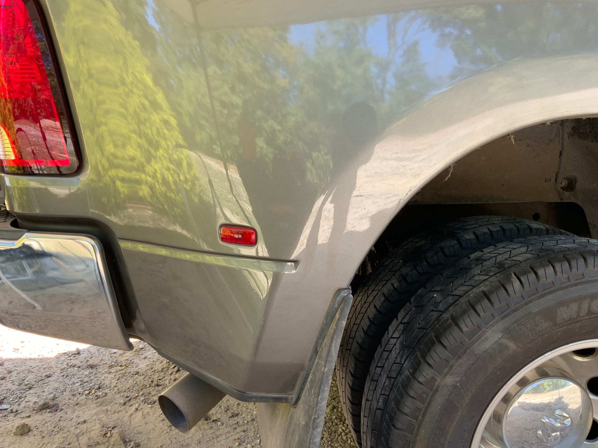 Close-up of the rear corner of a silver pickup truck showing part of the rear light, tire, and mud flap with a dirt ground and outdoor scenery reflected on the truck's surface.