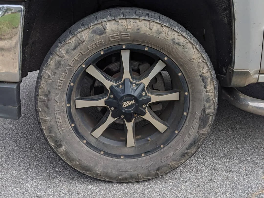 Close-up of a dirty off-road tire mounted on a black and gray alloy rim on a vehicle, with a dirt and gravel surface.