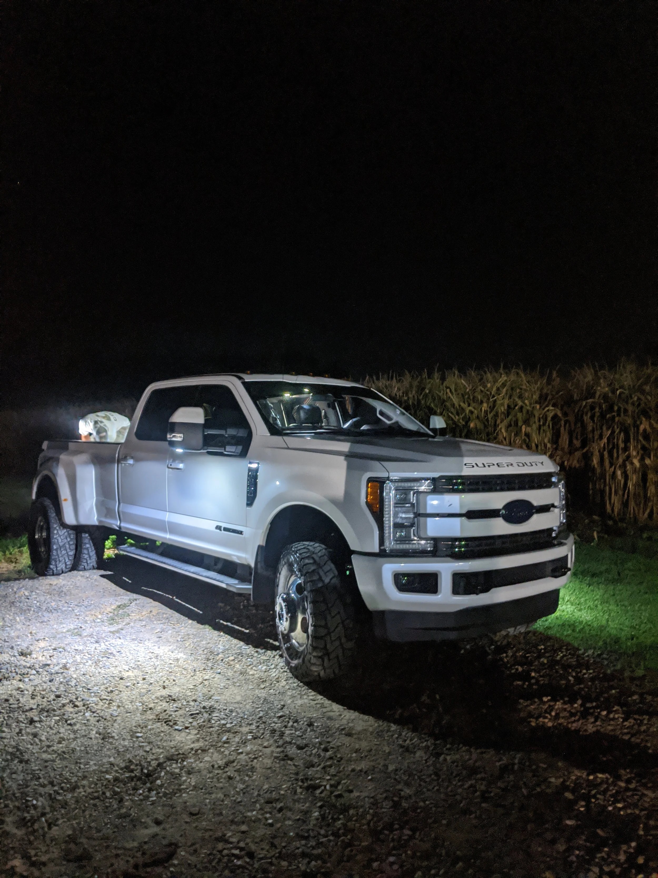 White Ford Super Duty pickup truck parked outdoors at night on a gravel surface, with cornfields in the background and headlights illuminating the scene.