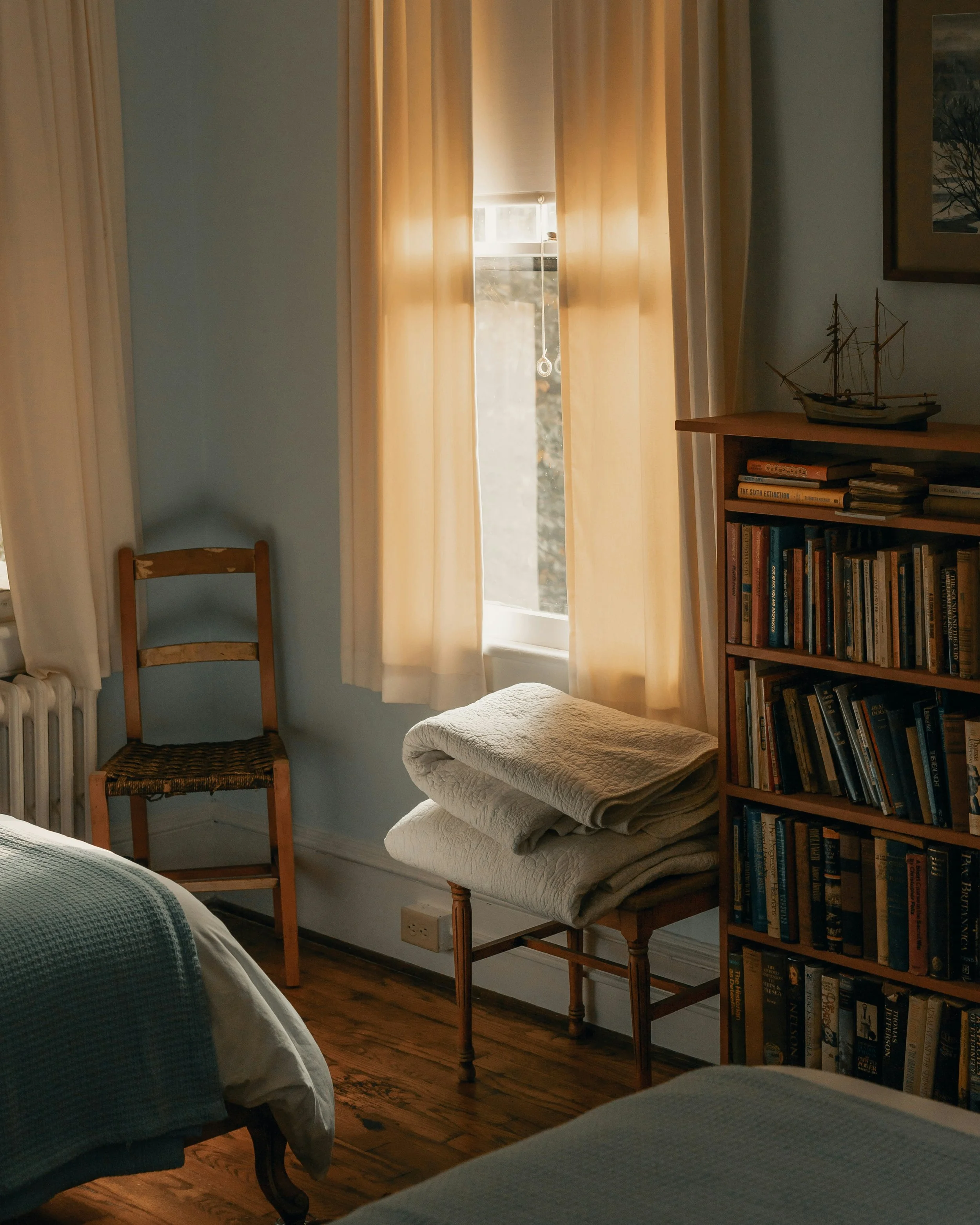 A cozy bedroom corner with a window covered by sheer curtains, a wooden chair with a woven seat, a small table stacked with folded towels, a bookshelf filled with books, a model ship on top of the bookshelf, and warm sunlight streaming through the window.