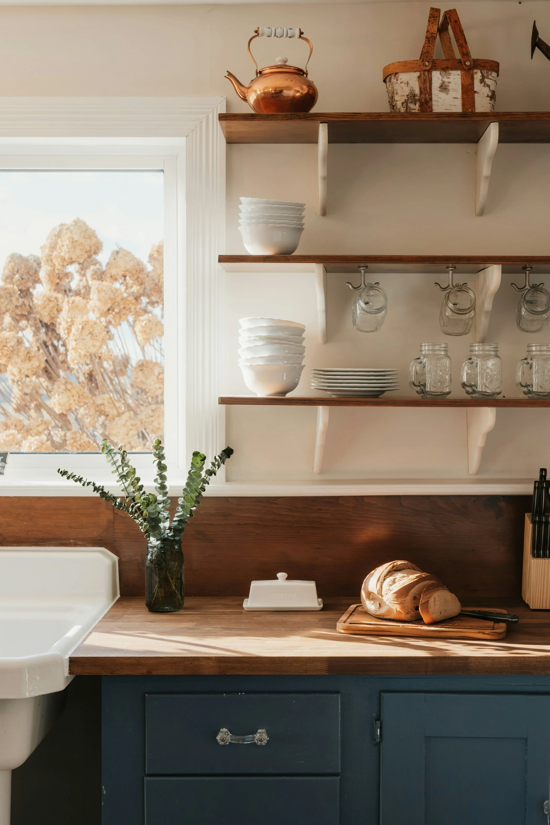 Kitchen with open wooden shelves holding bowls, plates, and jars, sunlight coming through a window with trees outside, a vase with greenery on a wooden countertop, a loaf of bread on a cutting board, and blue cabinetry.