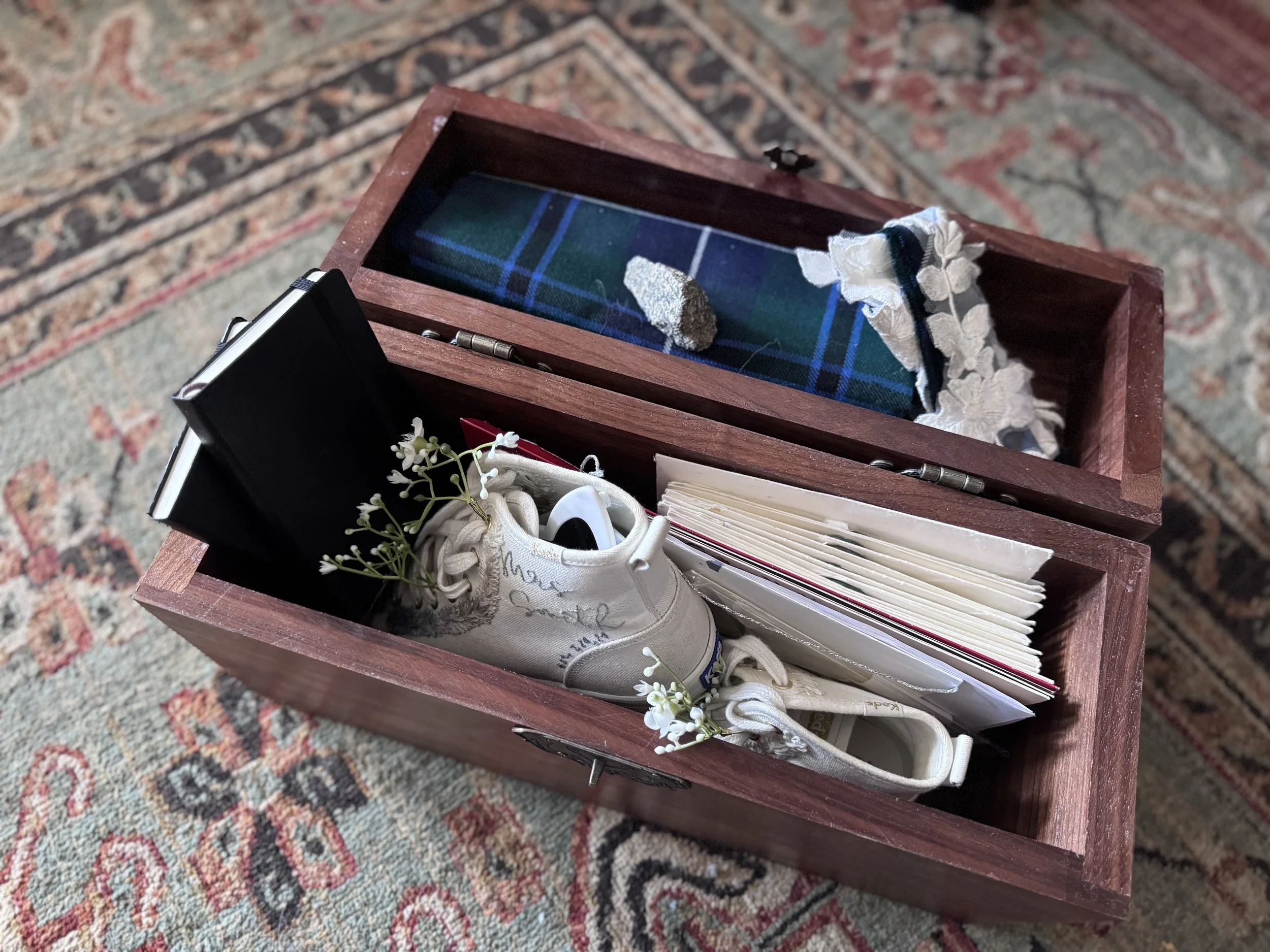 Open wooden keepsake box holding embroidered white sneakers, a folded tartan, a rock, lace, black vow books, cards, and small white flowers