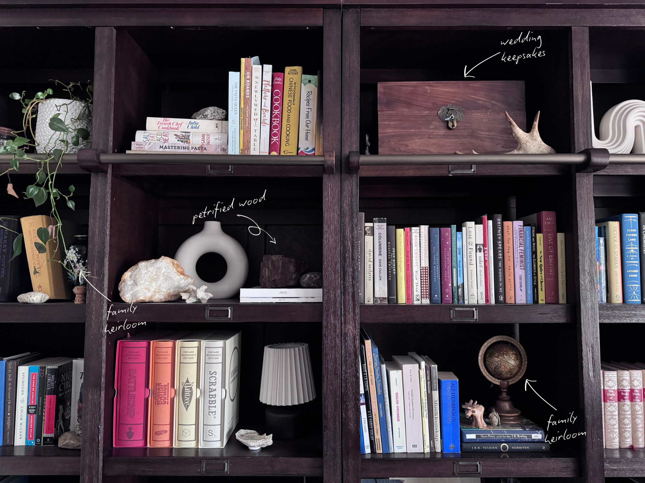 Dark wood bookshelves with cookbooks, novels, and board games, with handwritten labels pointing out a wooden box of wedding keepsakes, a piece of petrified wood, and two family heirlooms, including a small antique globe