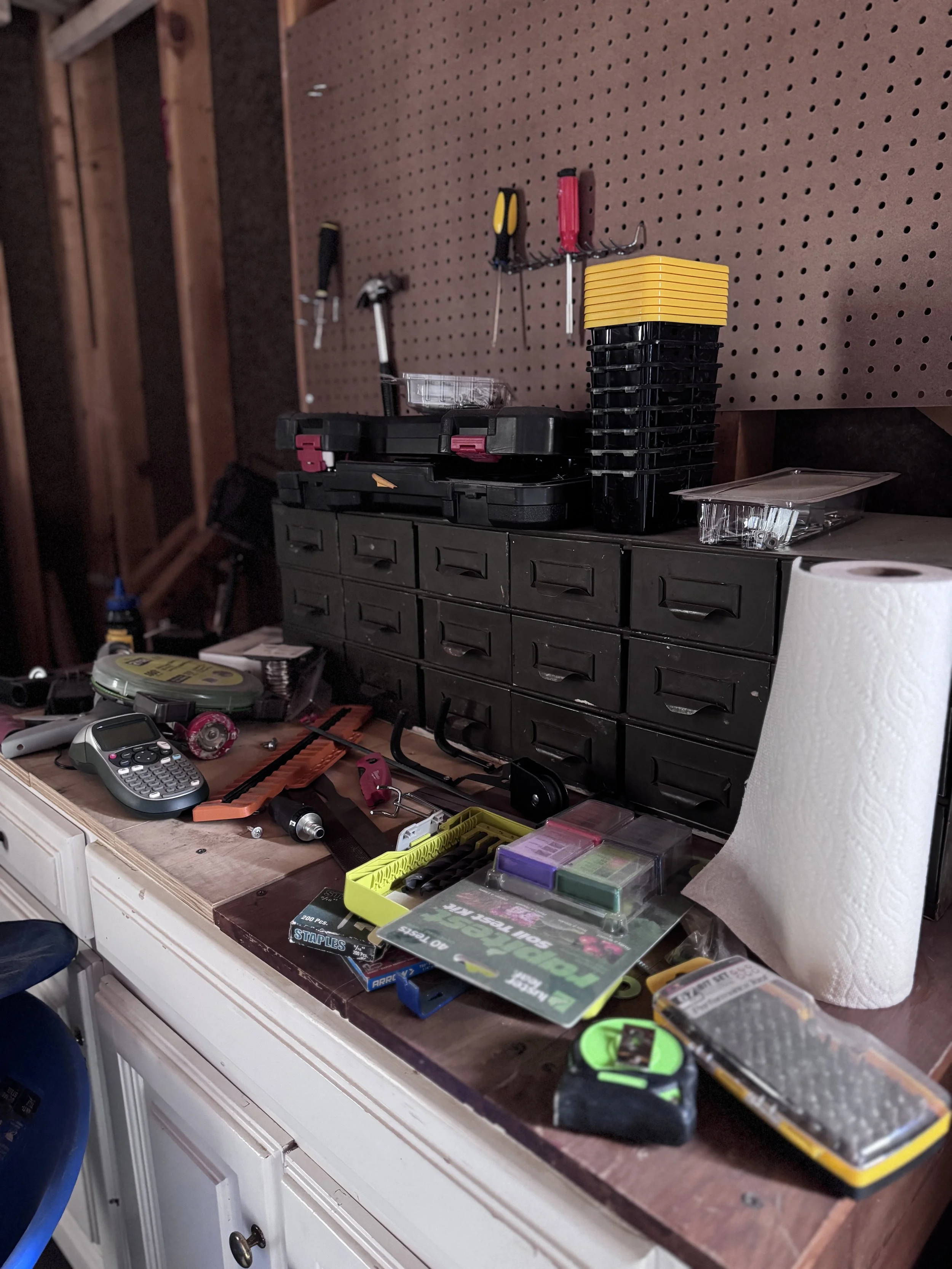Garage workbench covered in loose hardware, staples, drill bits, a tape measure, and a label maker, in front of a pegboard and a metal drawer organizer