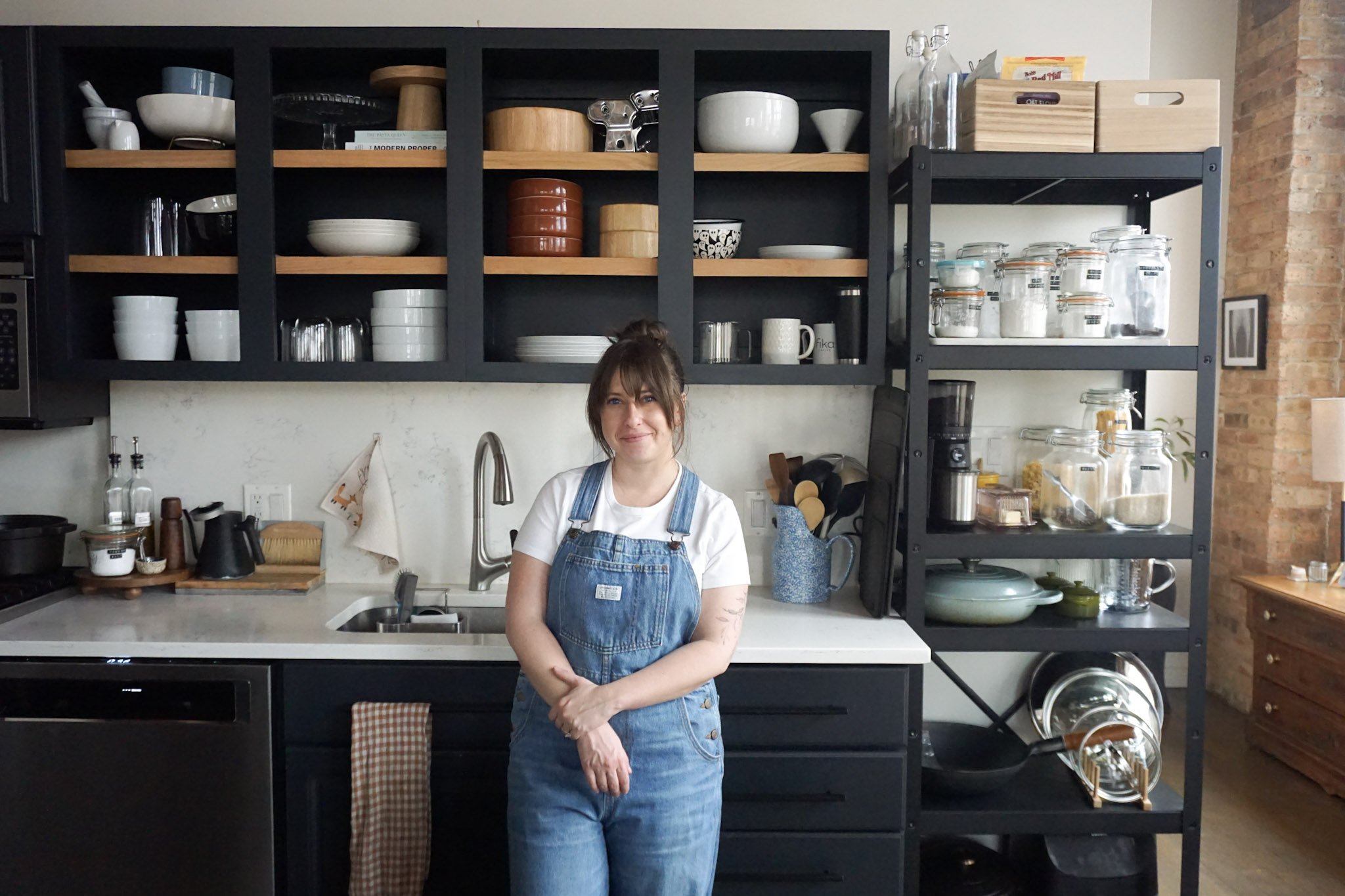 Woman in denim overalls standing in a modern kitchen with open shelving, dishes, glass jars, and cooking utensils.