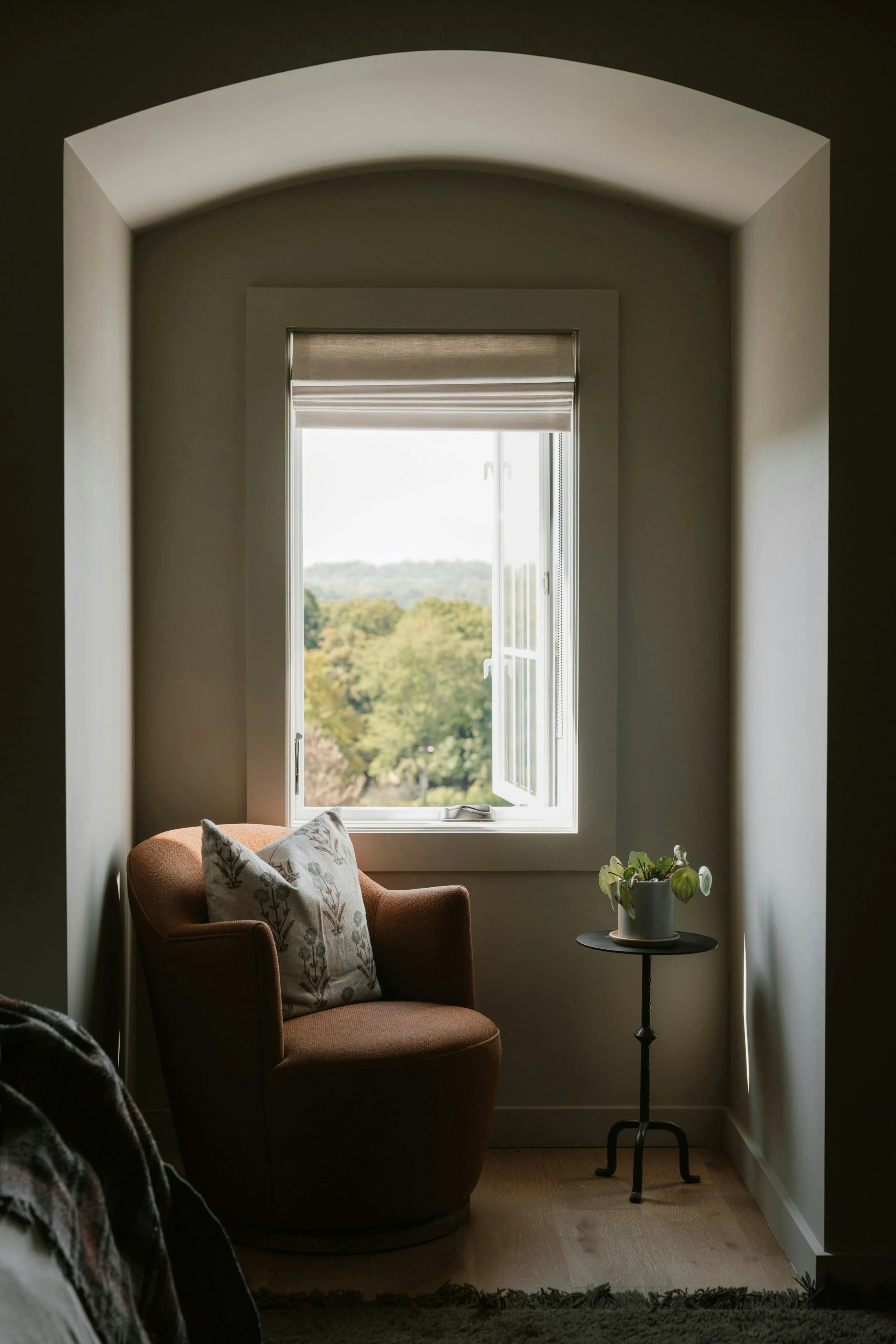 Cozy reading nook with a brown armchair, decorative pillow, and a small black side table with a potted plant, next to an open window with a view of trees and sky.