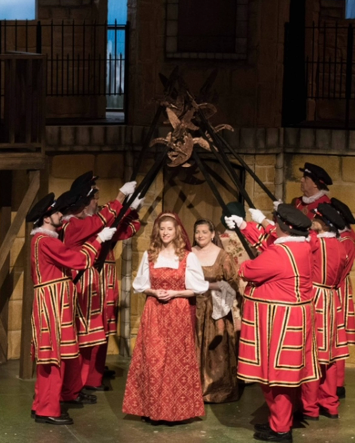 Actors on stage performing a scene from a play with medieval or renaissance costumes, including women in long dresses and men in red uniforms with black hats, in front of a backdrop depicting a stone building and a set piece resembling a gallows or a