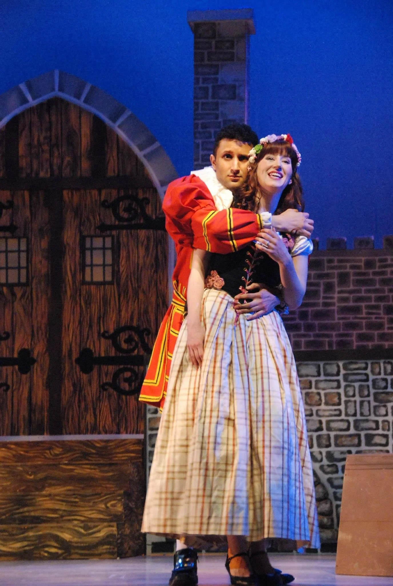Two actors performing on stage in costumes, with a backdrop resembling a house with a wooden door and stone chimney, during a theatrical play.