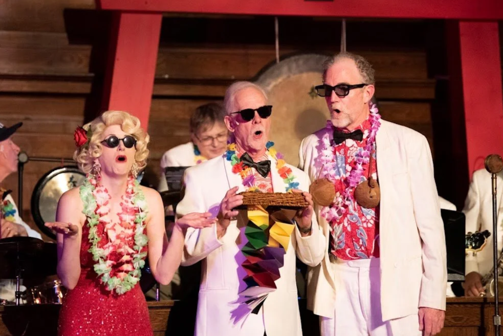 Three older adults singing on stage in tropical-themed party costumes, with a band playing in the background.