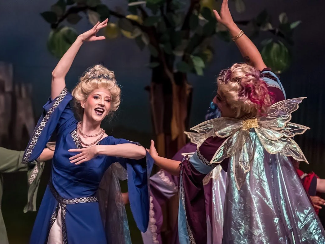 Two women dressed as fairies or magical characters dancing happily on stage with a backdrop of trees and other performers.