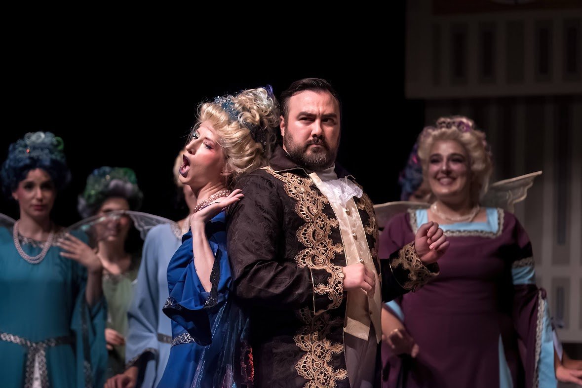 Scene from a theatrical play featuring a man in elaborate historical costume and women dressed as fairies or spirits with wings and colorful costumes.
