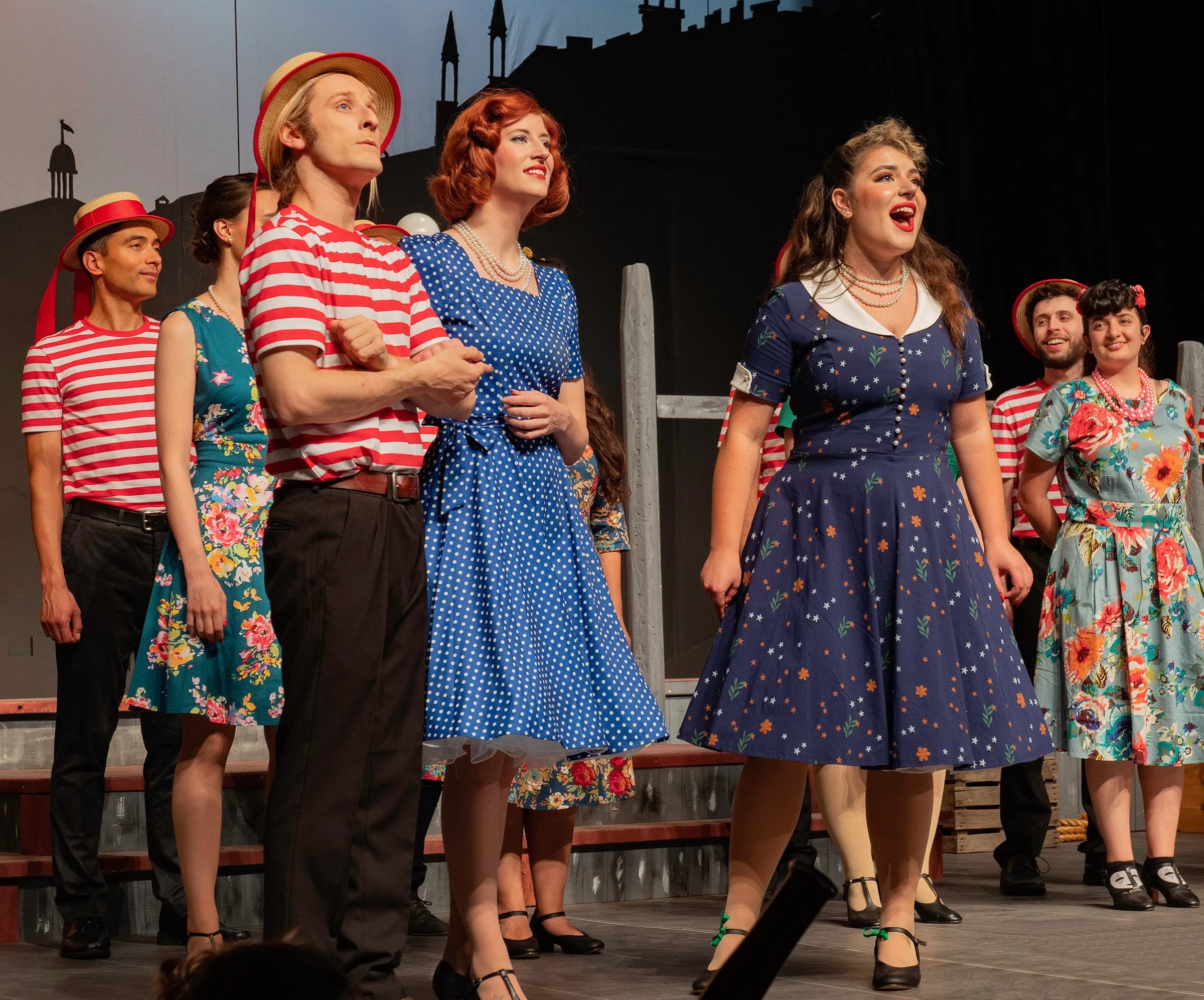 Stage performance with actors in vintage 1950s-style dresses and striped shirts, some wearing hats, standing in a row during a theatrical show.