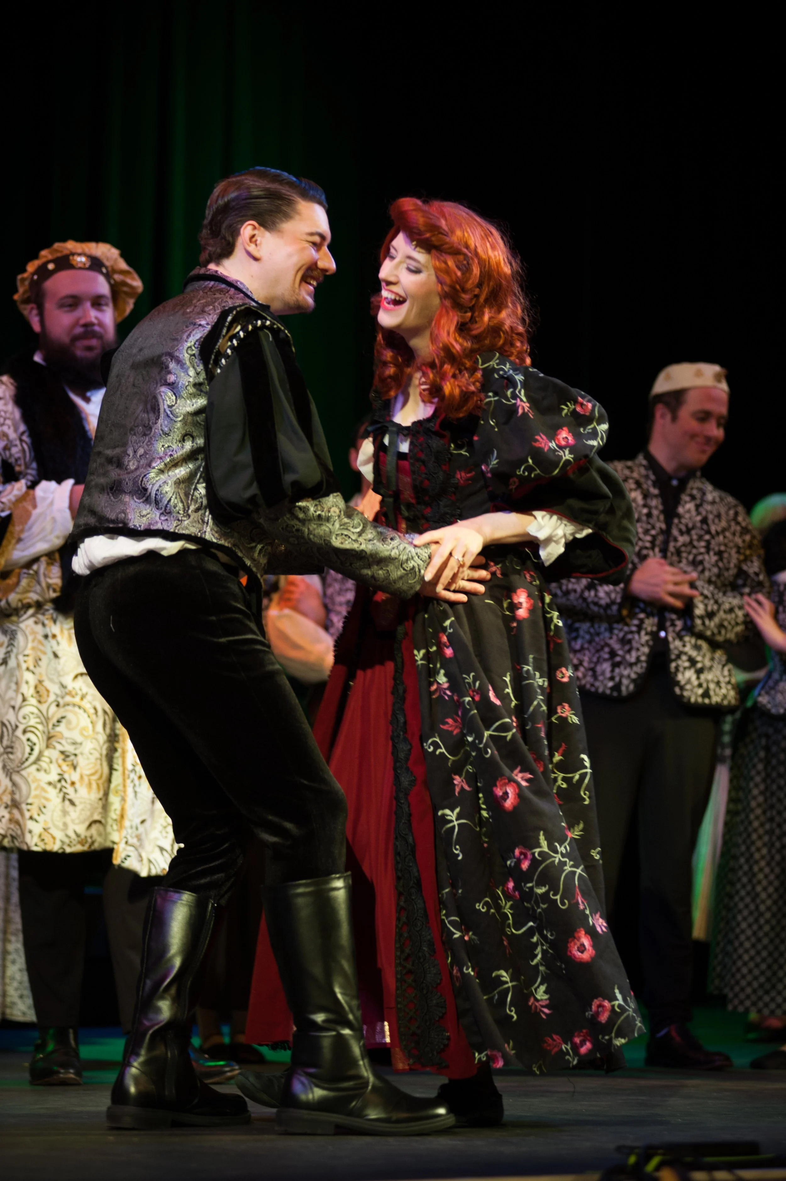 Actors on stage performing a scene dressed in historical costumes, with a man and woman smiling and holding hands, surrounded by other performers.
