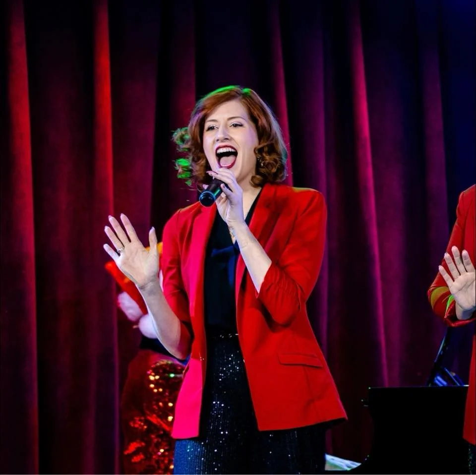 A woman singing on stage with a microphone, wearing a red blazer and dark sparkly pants, in front of purple curtains.