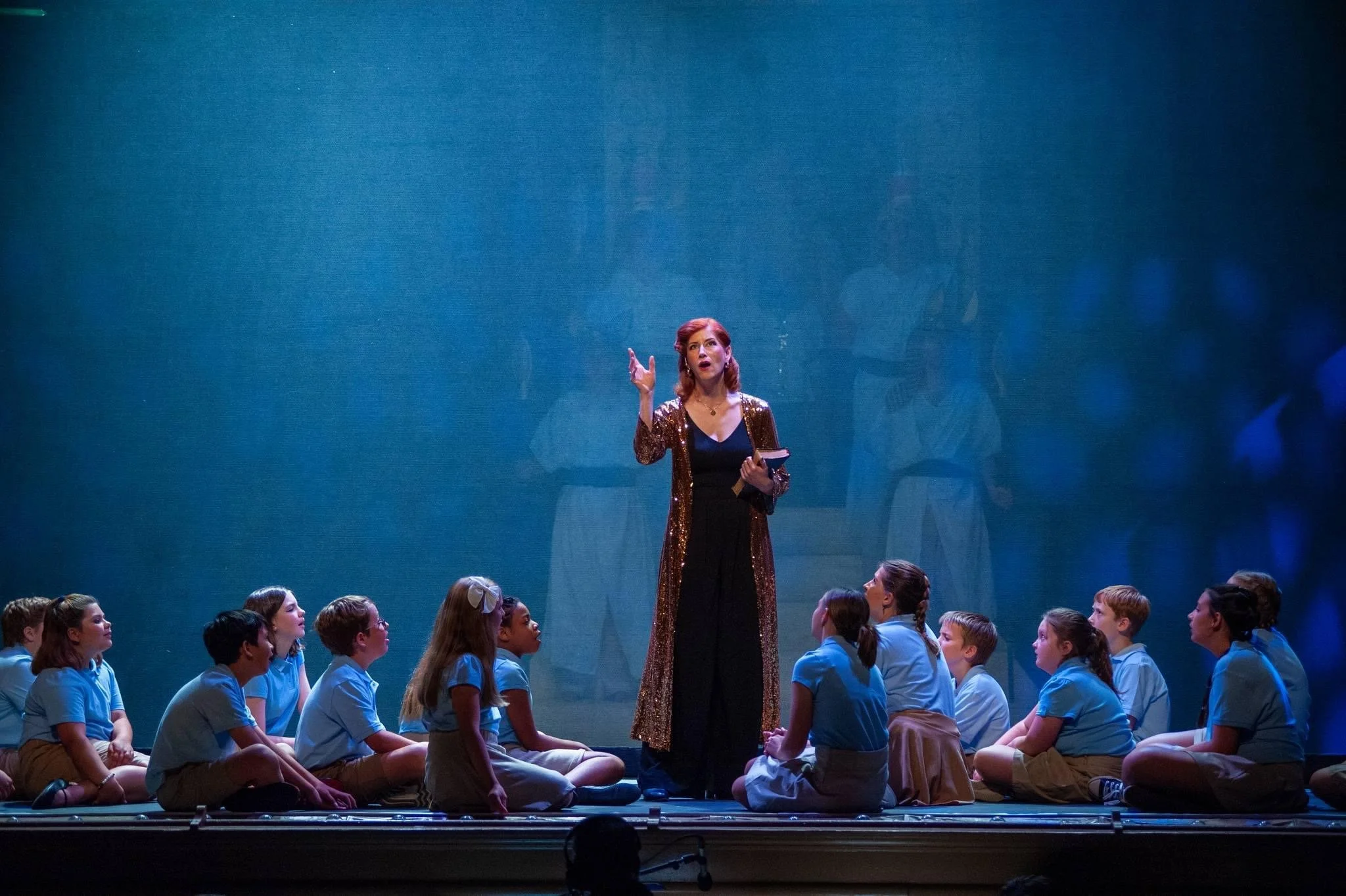 Stage scene with a woman singing or speaking, wearing a long sequined coat, surrounded by young people in blue shirts sitting cross-legged, with a blue backdrop.