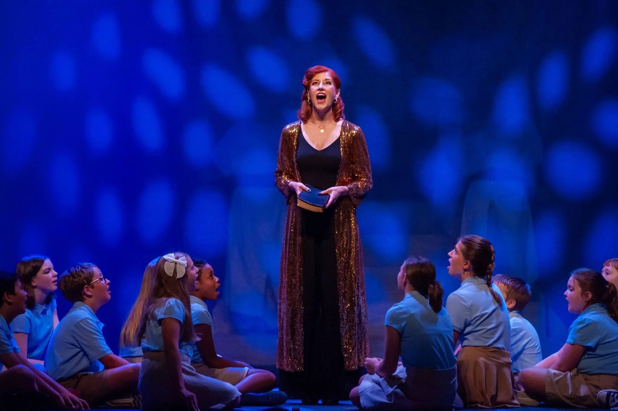 A theatrical performance scene with a woman standing at the center, speaking to children seated on the floor. The woman has red hair and is wearing a black dress and a long, sparkling, gold coat. The children are dressed in blue shirts and are lookin