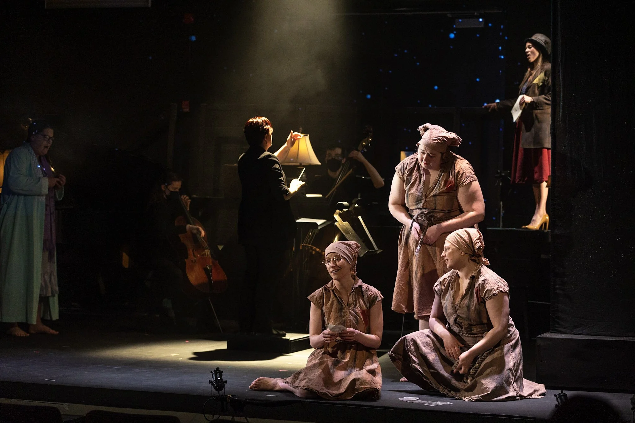 Theatrical stage performance with a group of women dressed in costumes resembling laundry, some kneeling and some standing, while musicians and a conductor are in the background.