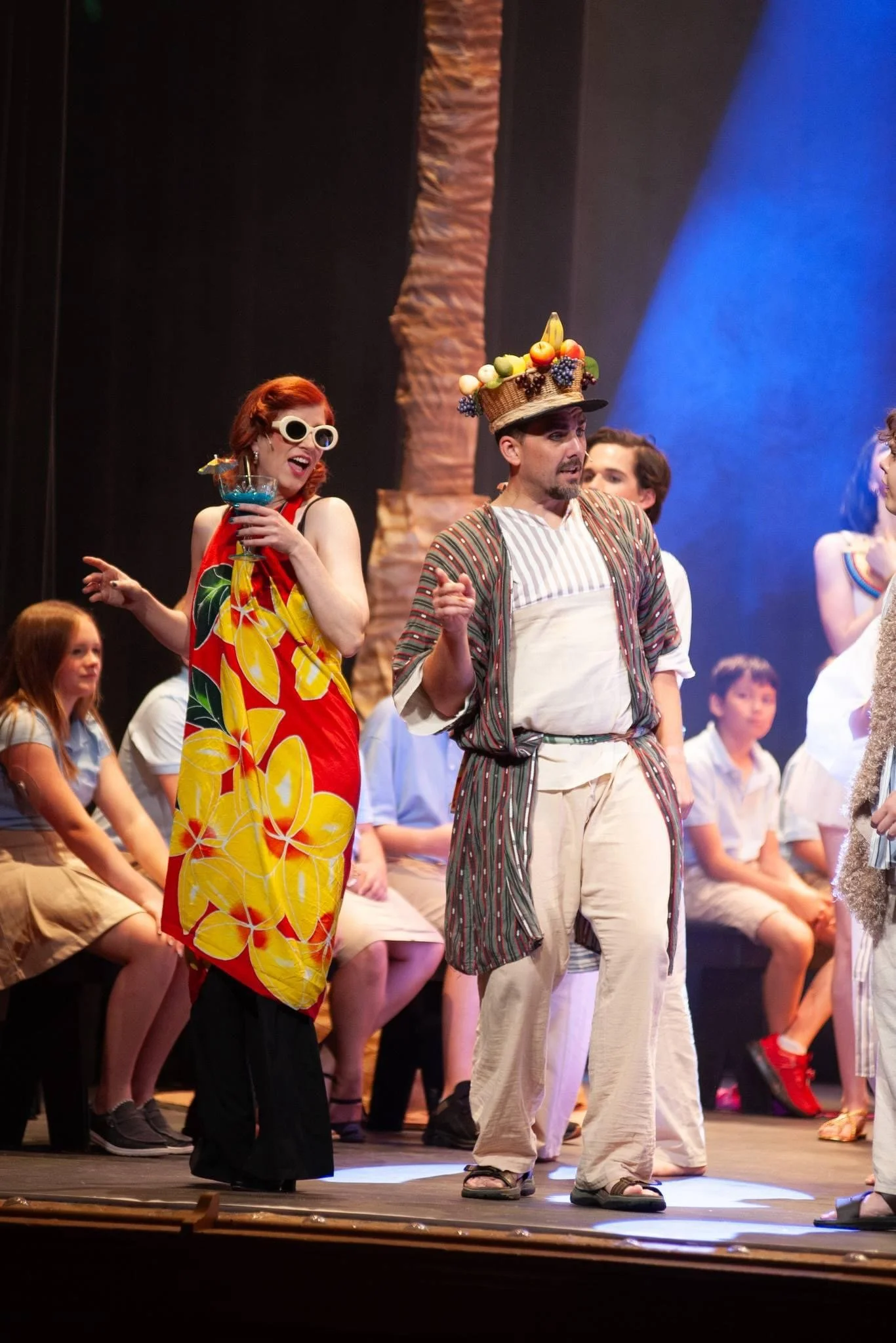 The image shows a theatrical stage with actors, including a woman with red hair, white sunglasses, and colorful clothing, and a man wearing a large fruit basket hat and casual attire, performing in a scene with an audience of children in the backgrou