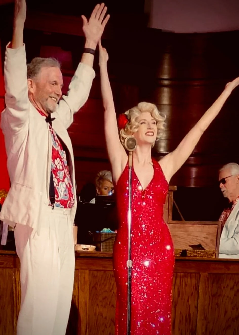 A man and a woman dressed in vintage clothing celebrating on stage with their hands raised, during a lively event.