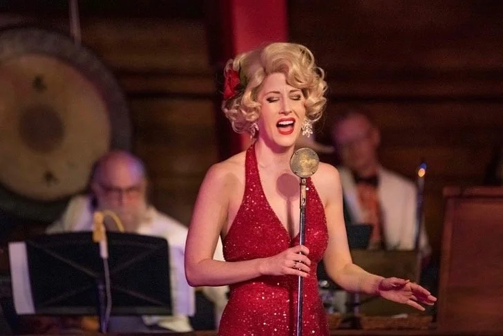 A woman singing passionately in a vintage-style red dress, with a band playing in the background.