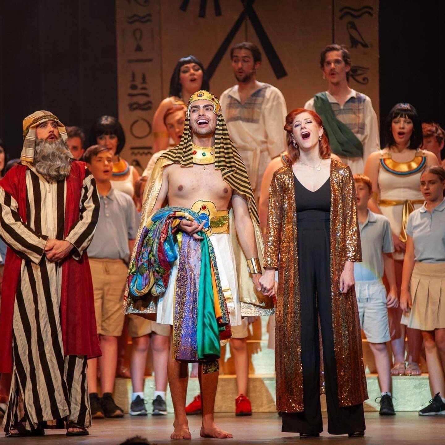 Group of performers on stage during a theatrical production. One man dressed as an Egyptian Pharaoh in the center, holding hands with a woman in a gold and black outfit, while others sing or speak in the background.
