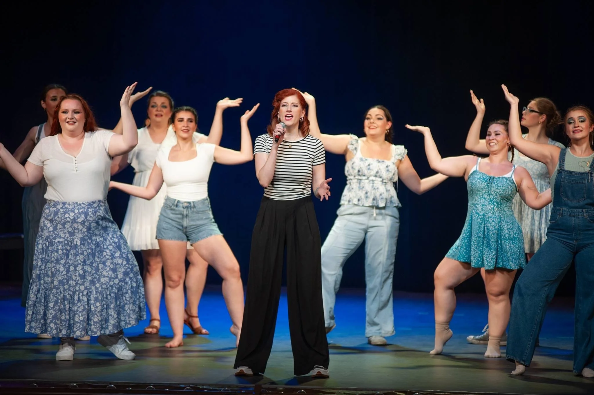 A group of women on stage, with one woman in the center singing into a microphone, dressed in casual summer clothes with a dark background.