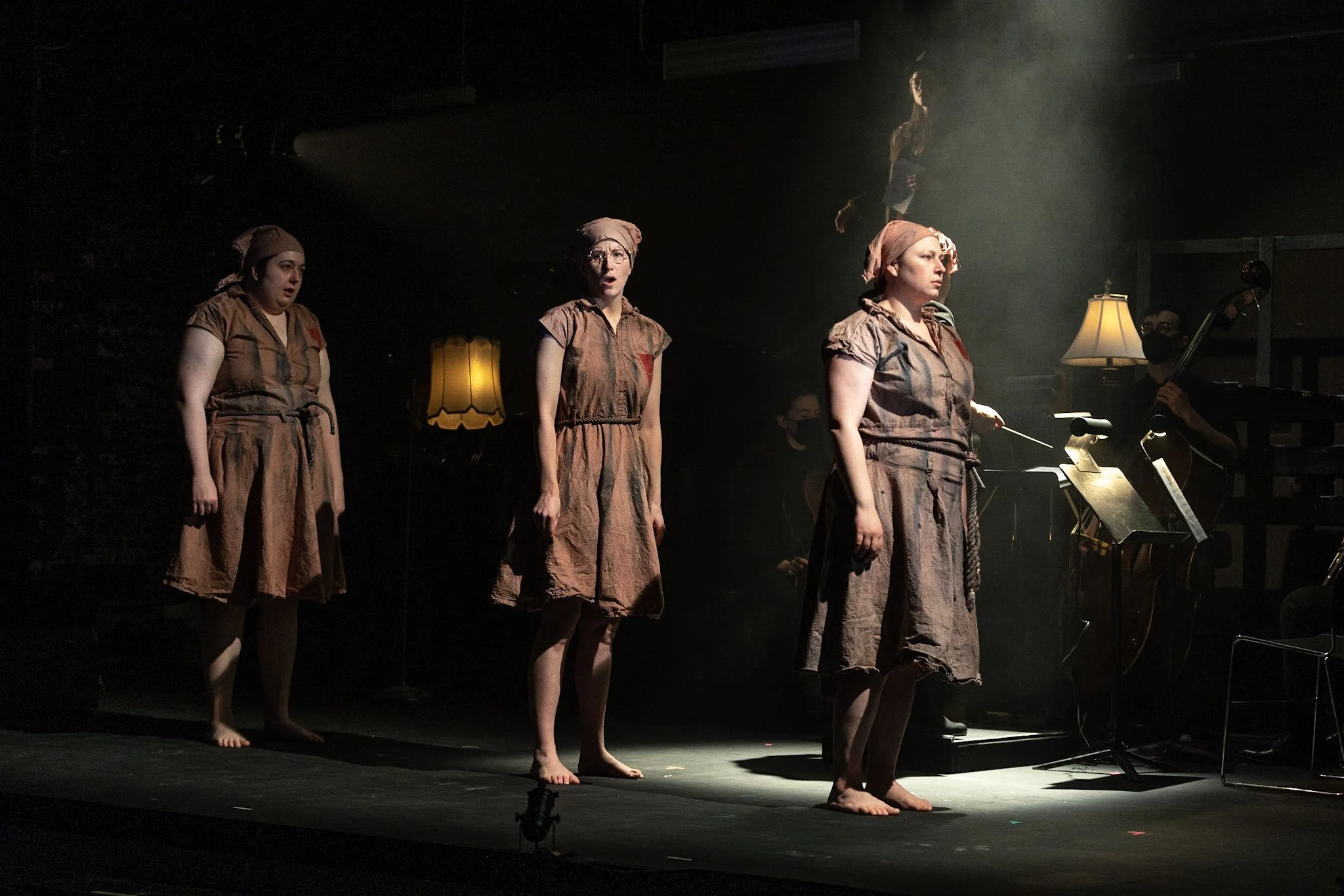 Three women dressed in brown costumes with head coverings, standing barefoot on stage, with a person playing a guitar in the background, during a theatrical performance.