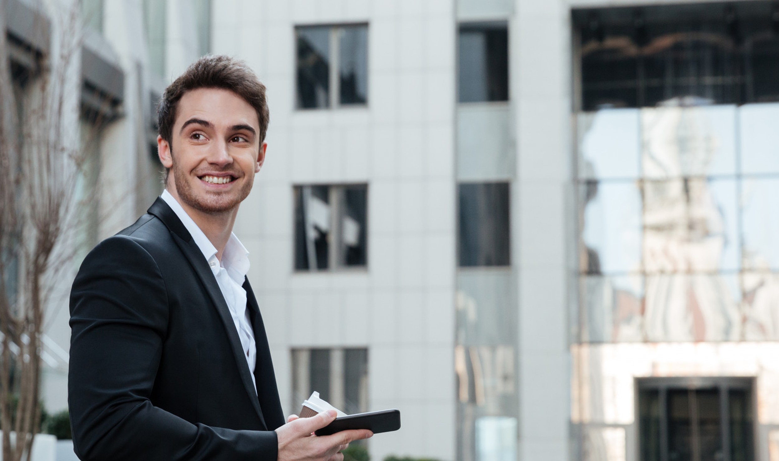 A young man in a black suit smiling while holding a coffee cup and a smartphone outside a modern building.