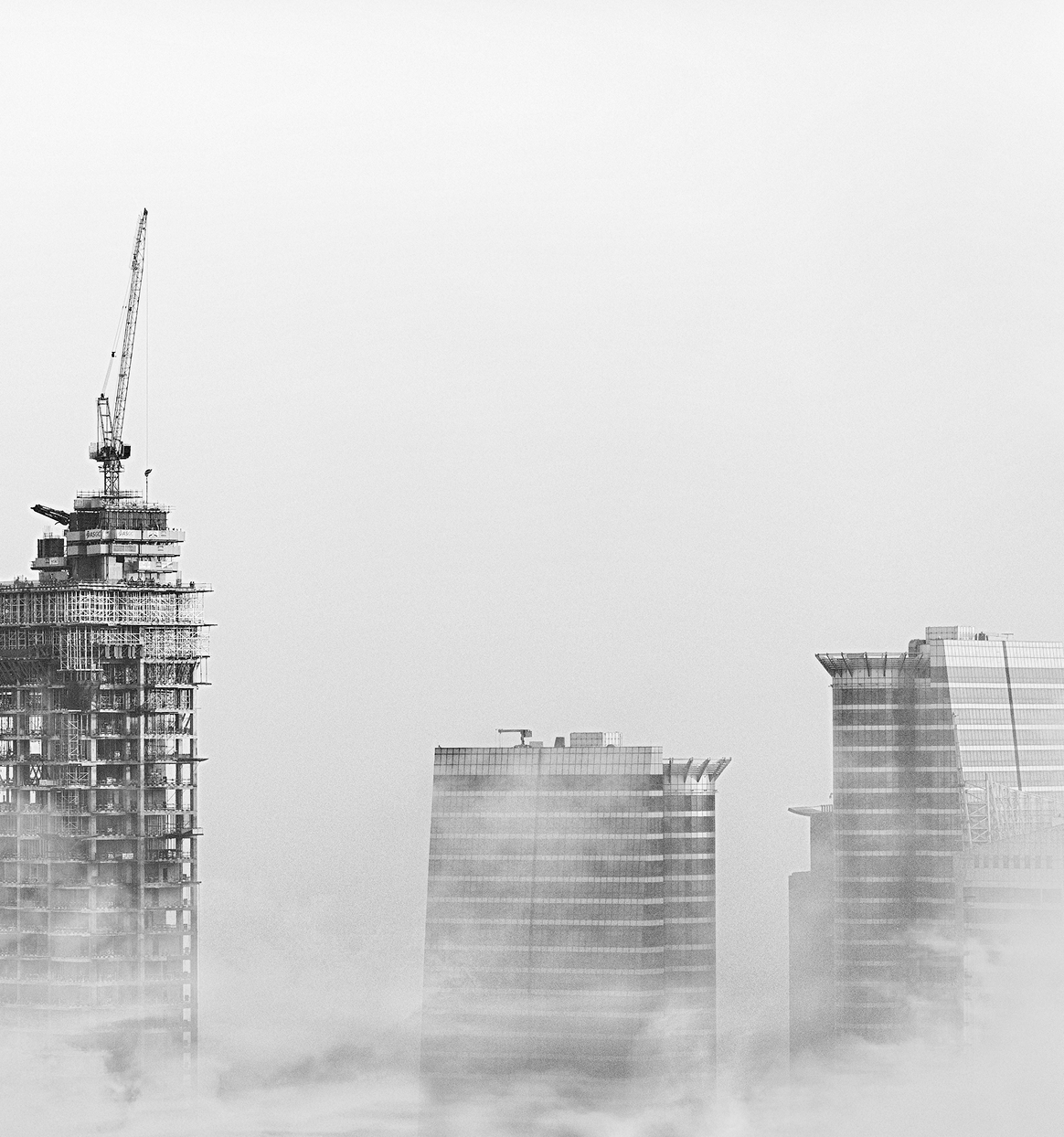 Black and white photo of three skyscrapers partially obscured by fog, with one under construction and a crane on top.