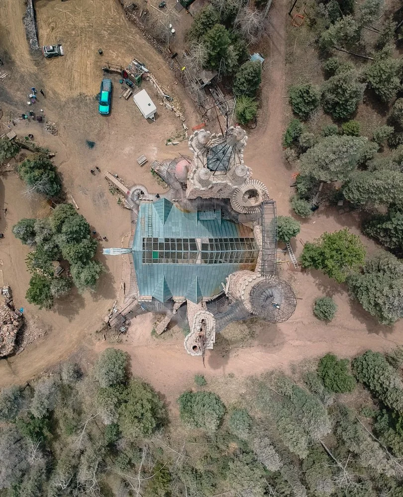 An aerial view of a colorful castle or building with a glass roof, surrounded by trees and dirt paths, with construction materials or equipment nearby.
