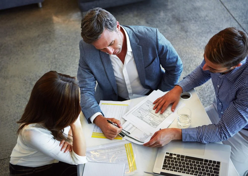 Three professionals discussing documents around a table with a laptop, water, and charts.