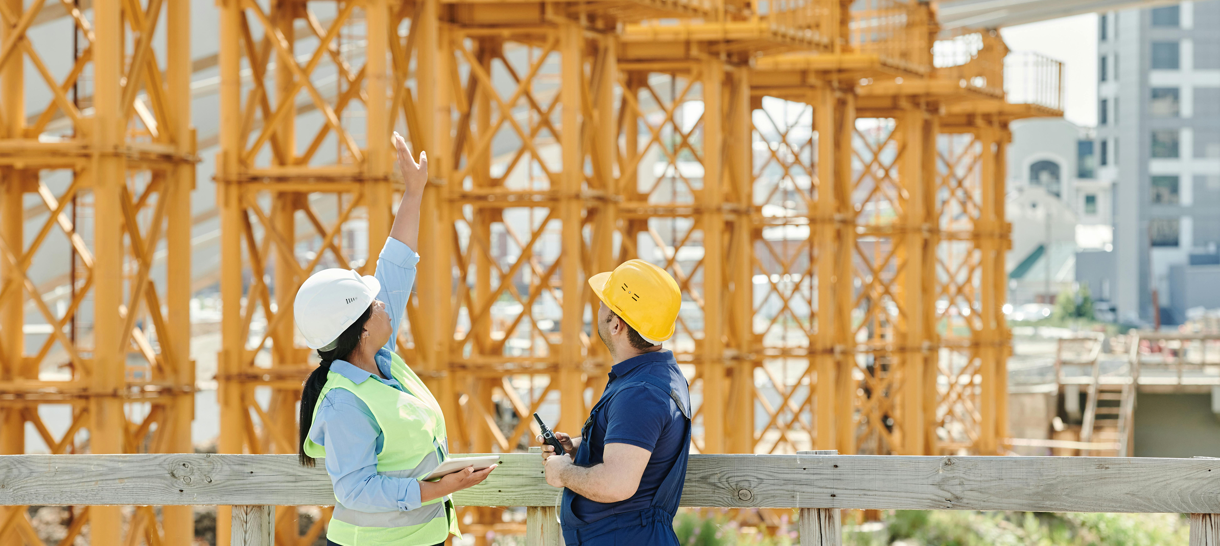 Two construction workers, a woman and a man, wearing safety helmets and vests, are discussing on a construction site with orange structural framework and urban buildings in the background.