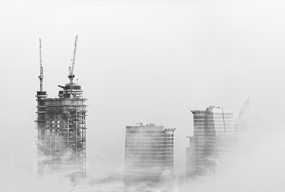 Skyline of tall skyscrapers emerging from fog with cranes on one building under construction.