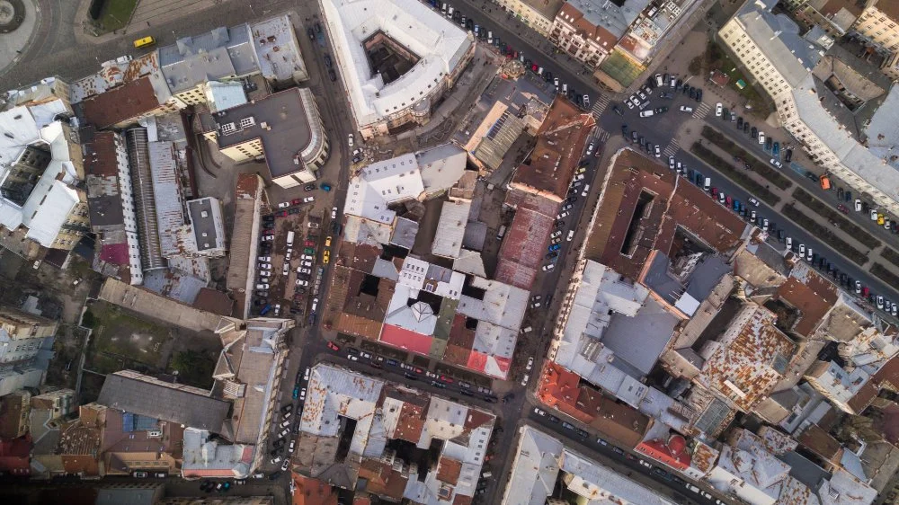Aerial view of tightly packed buildings and streets in an urban area, with cars parked along the roads and a small green space visible in the bottom left corner.