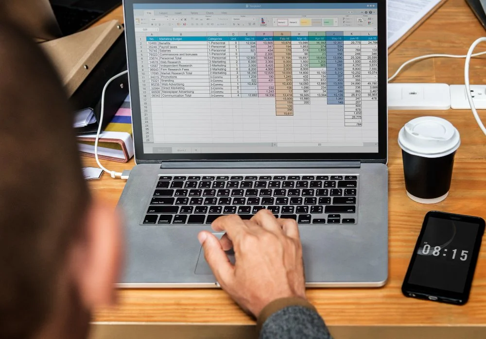 A person working on a laptop with an Excel spreadsheet, a smartphone displaying the time 08:15, a to-go coffee cup, and a power strip on a wooden desk.
