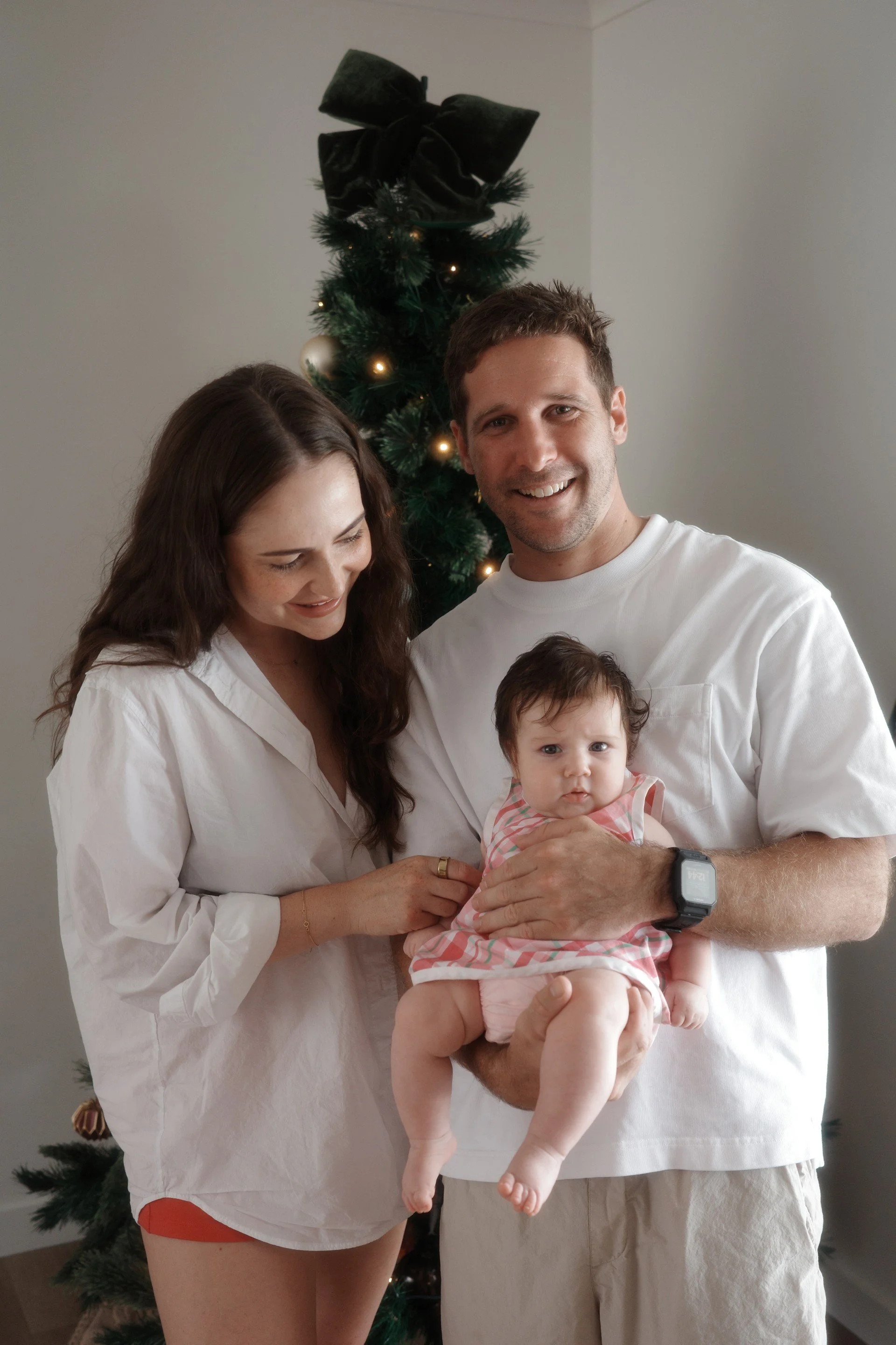 A smiling man holding a baby girl, standing in front of a decorated Christmas tree with a large black bow on top, in a room with plain walls.