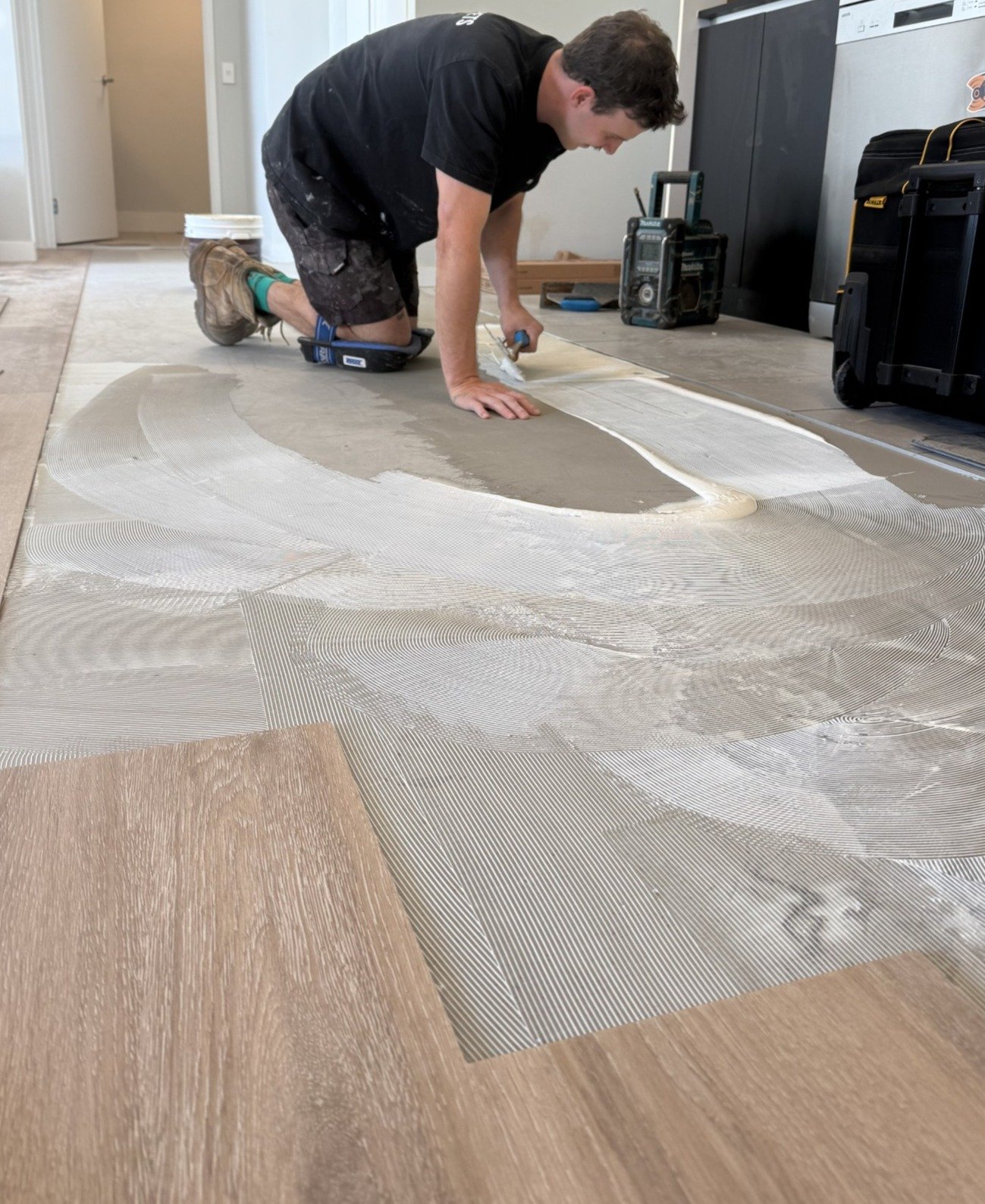 A person kneeling on the floor using a notched trowel to install a patterned tile floor. The person is wearing casual work clothes and a black t-shirt, surrounded by tools and equipment.