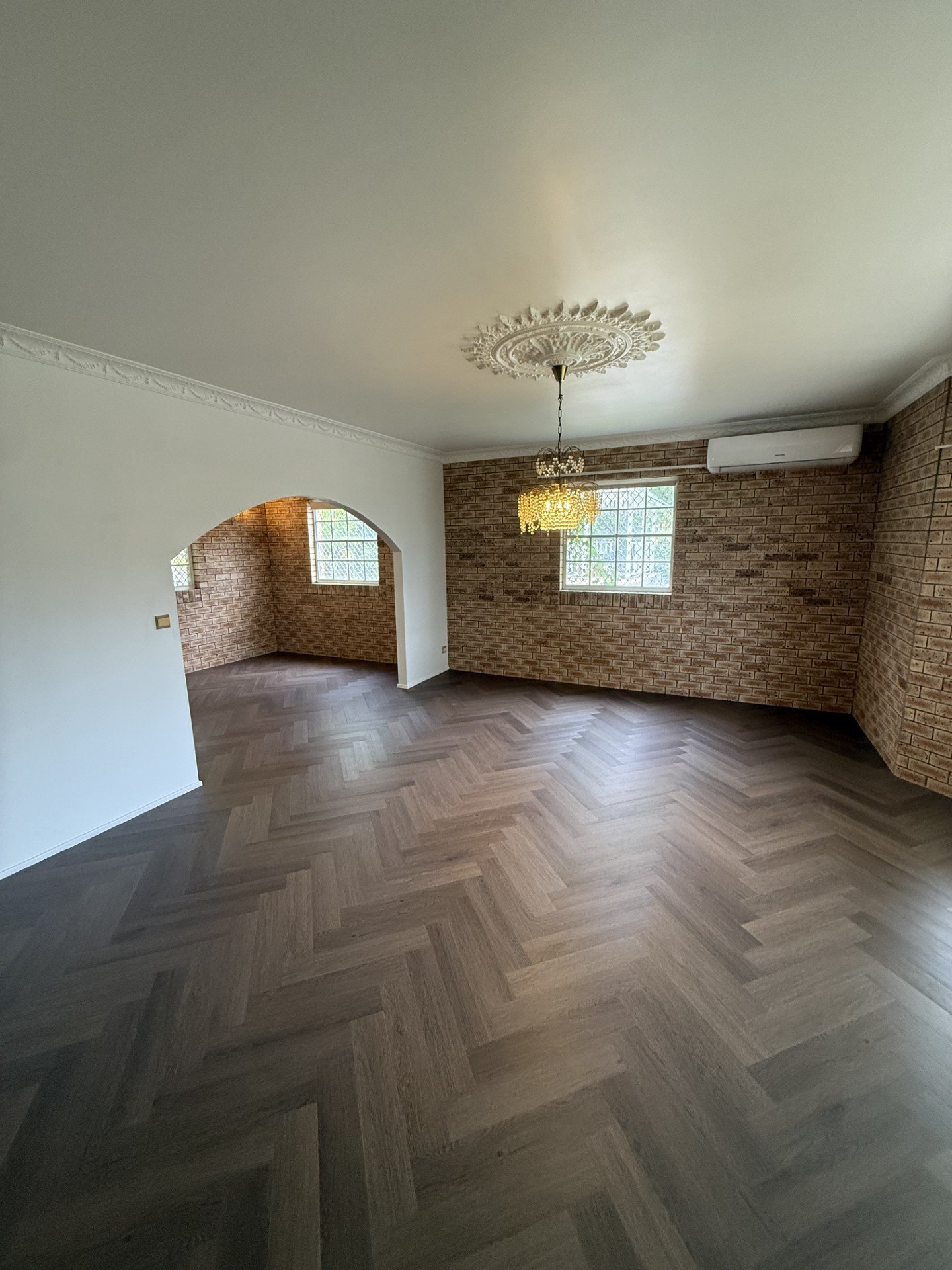 Living room with brick walls, hardwood herringbone floor, arched doorway, two windows, air conditioning unit, and a hanging chandelier.