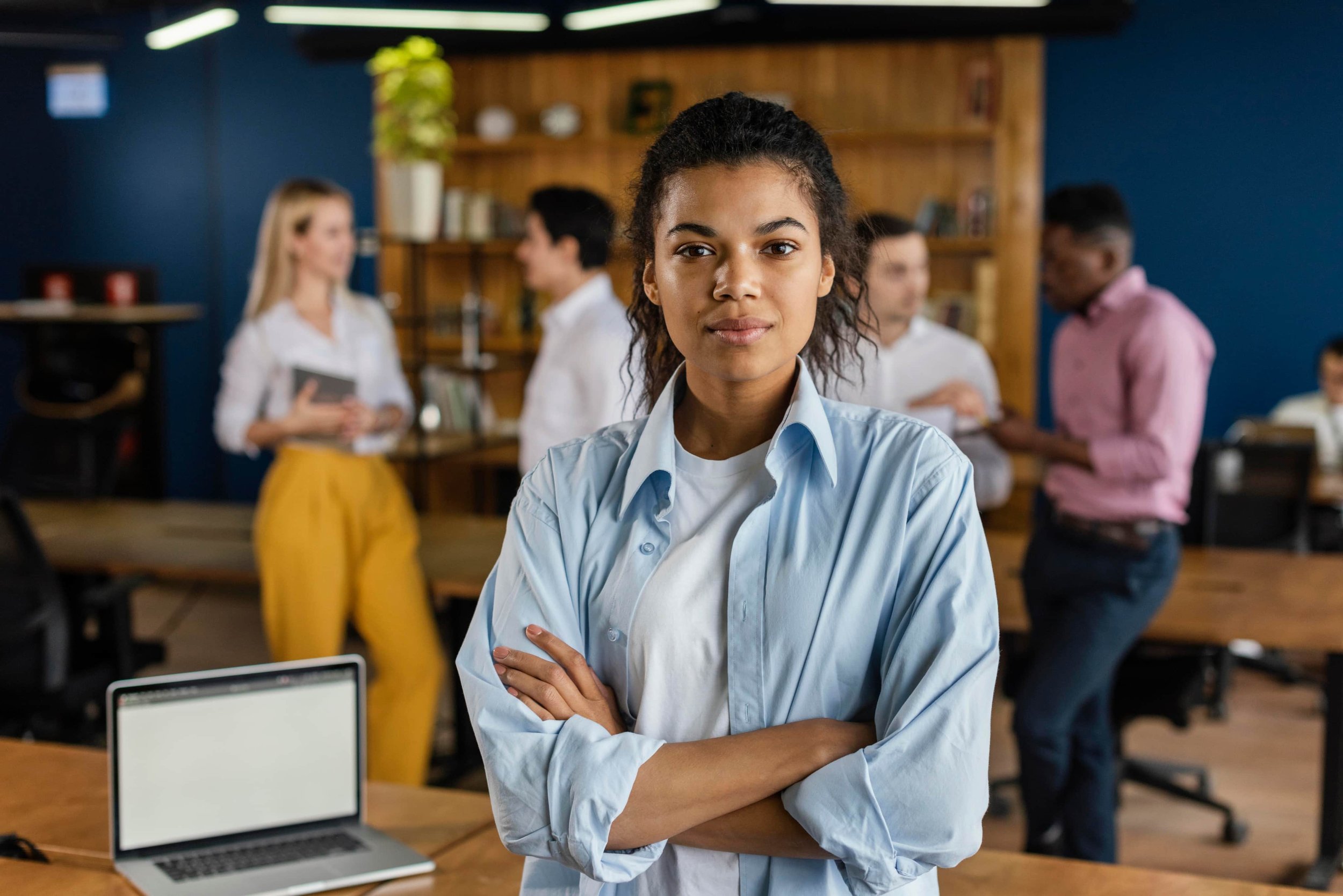 Young woman standing with arms crossed in a modern office space, with a laptop on the desk in front of her and colleagues in the background