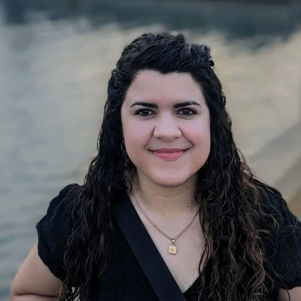 A woman with dark curly hair, smiling, wearing a black top and a gold necklace with a square pendant, standing outdoors near water.
