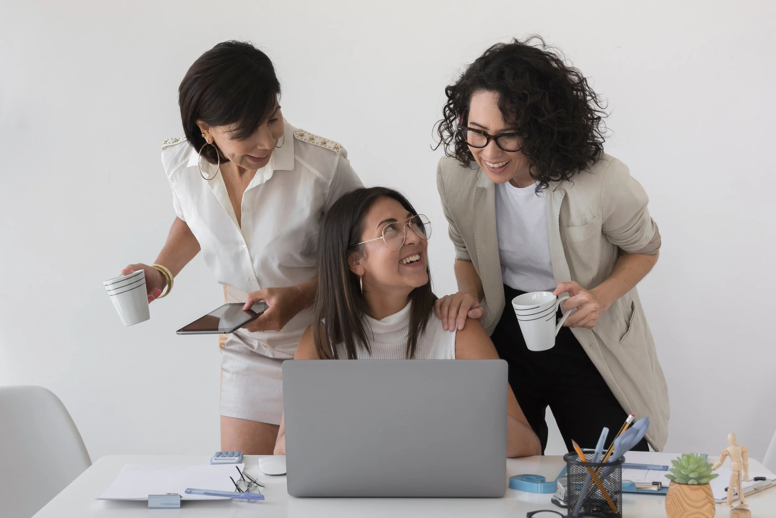 Three women are gathered around a desk with a laptop, smiling and talking. One woman sits, wearing glasses and a white sleeveless top, while the other two women stand, holding coffee mugs. The desk has office supplies and papers.