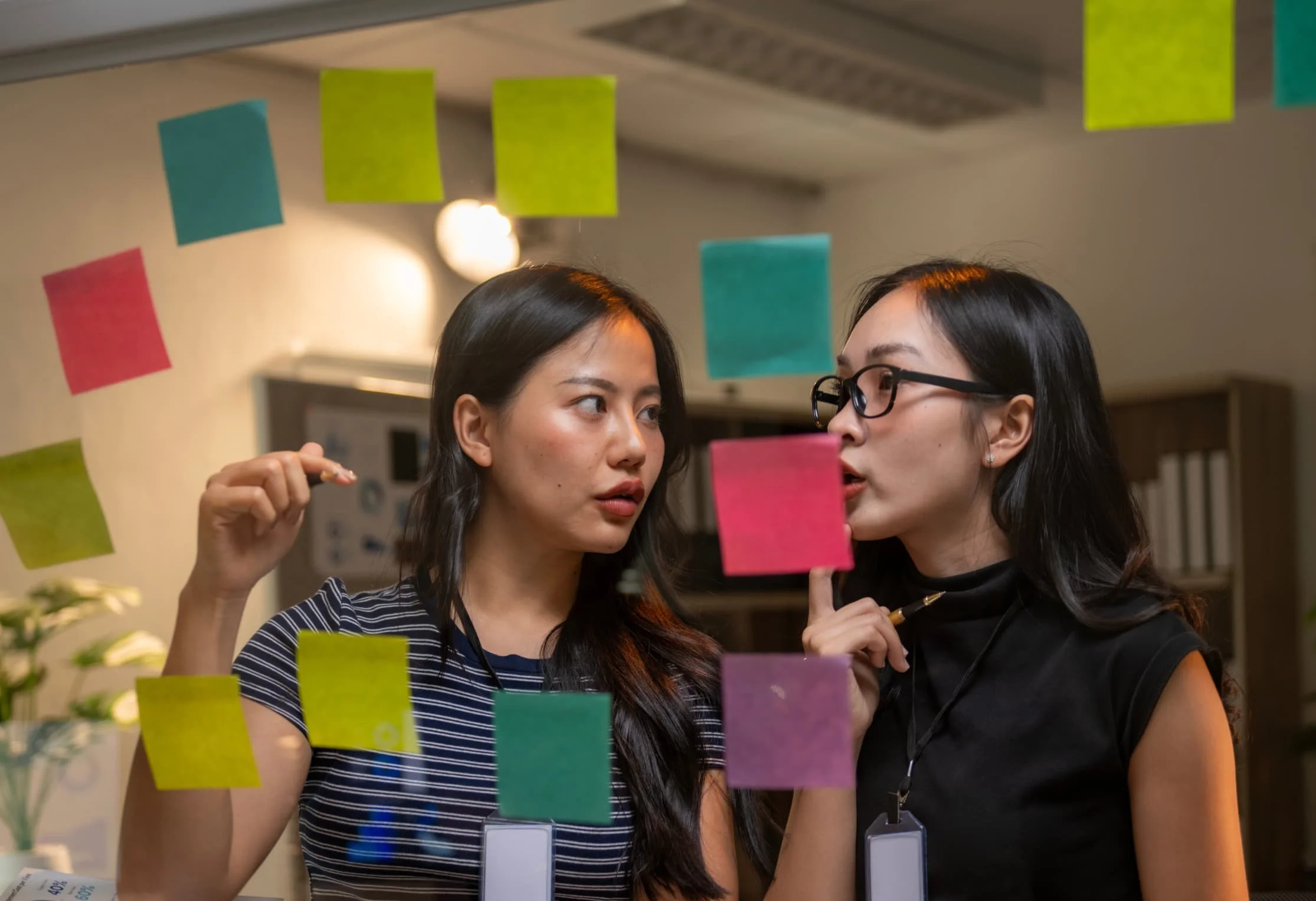 Two women are working together with sticky notes on a glass wall in an office setting.