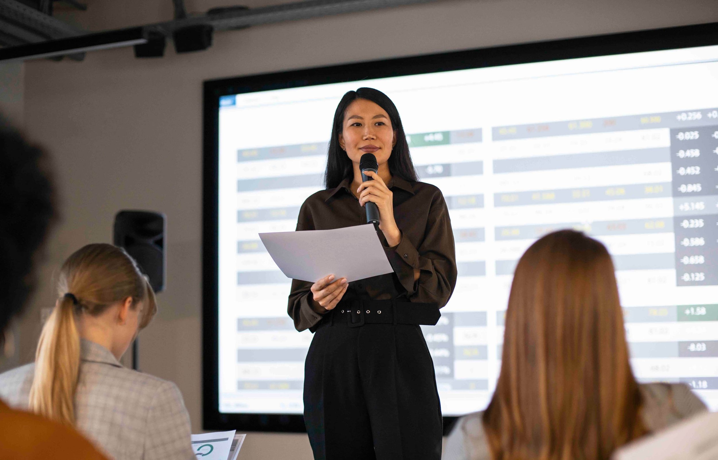 A woman holding a microphone and some papers giving a presentation in front of a large screen displaying financial data, with audience members watching.