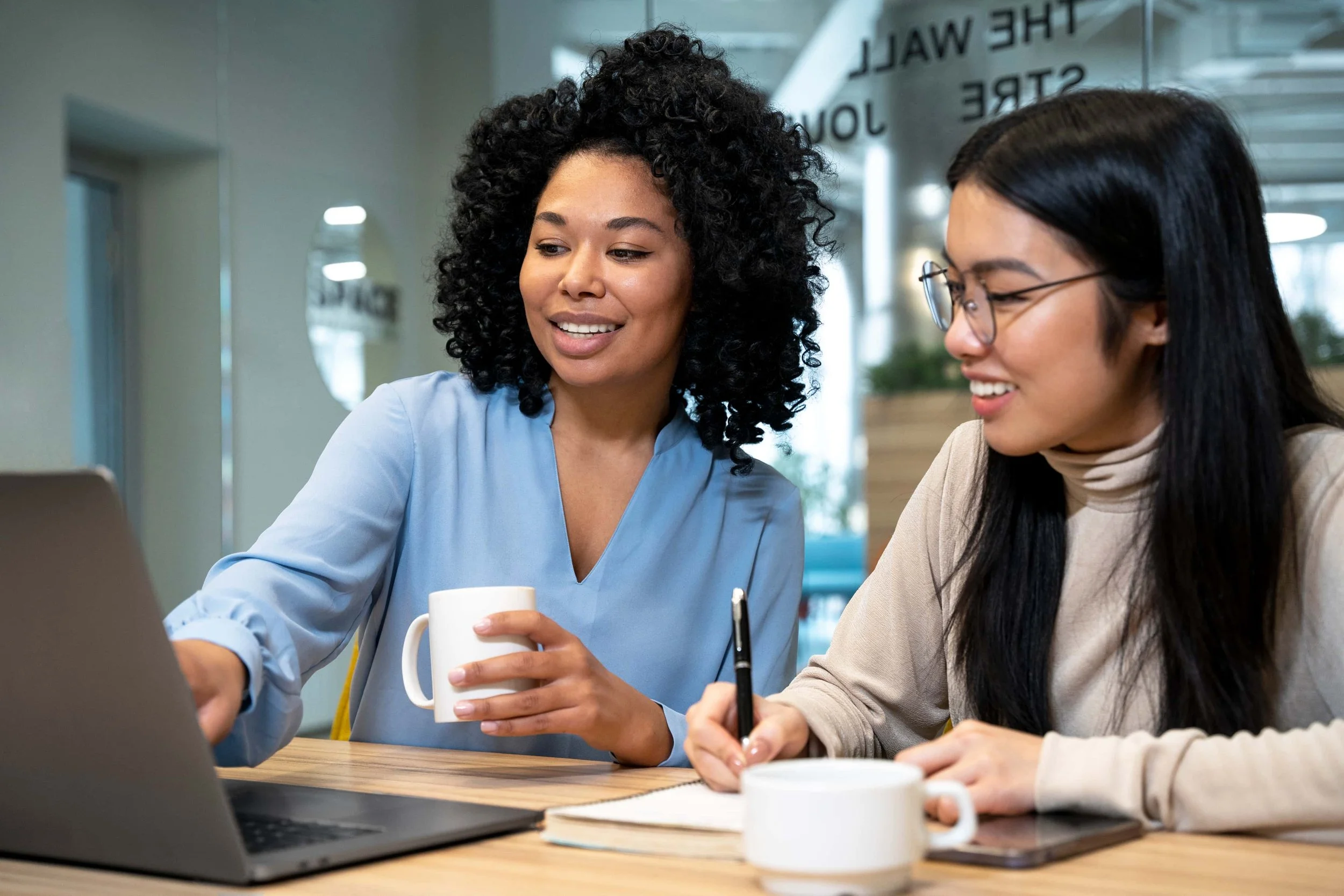 Two women sitting at a table with laptops and coffee mugs, collaborating in a office space.