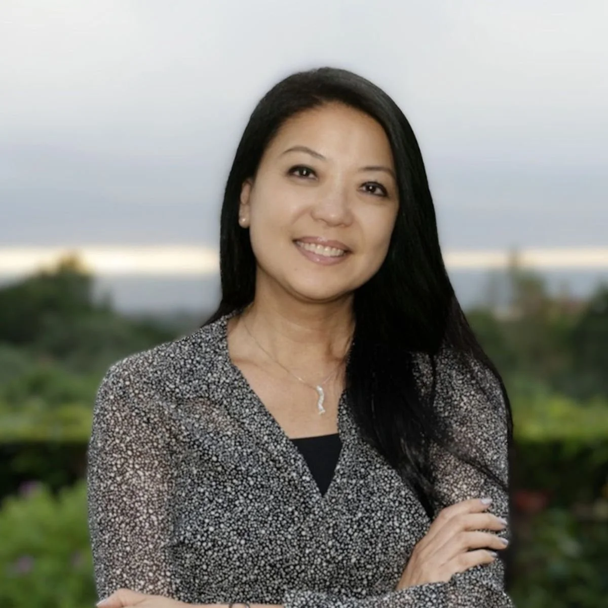 A woman with long black hair smiling outdoors with a blurred background of trees and sky.