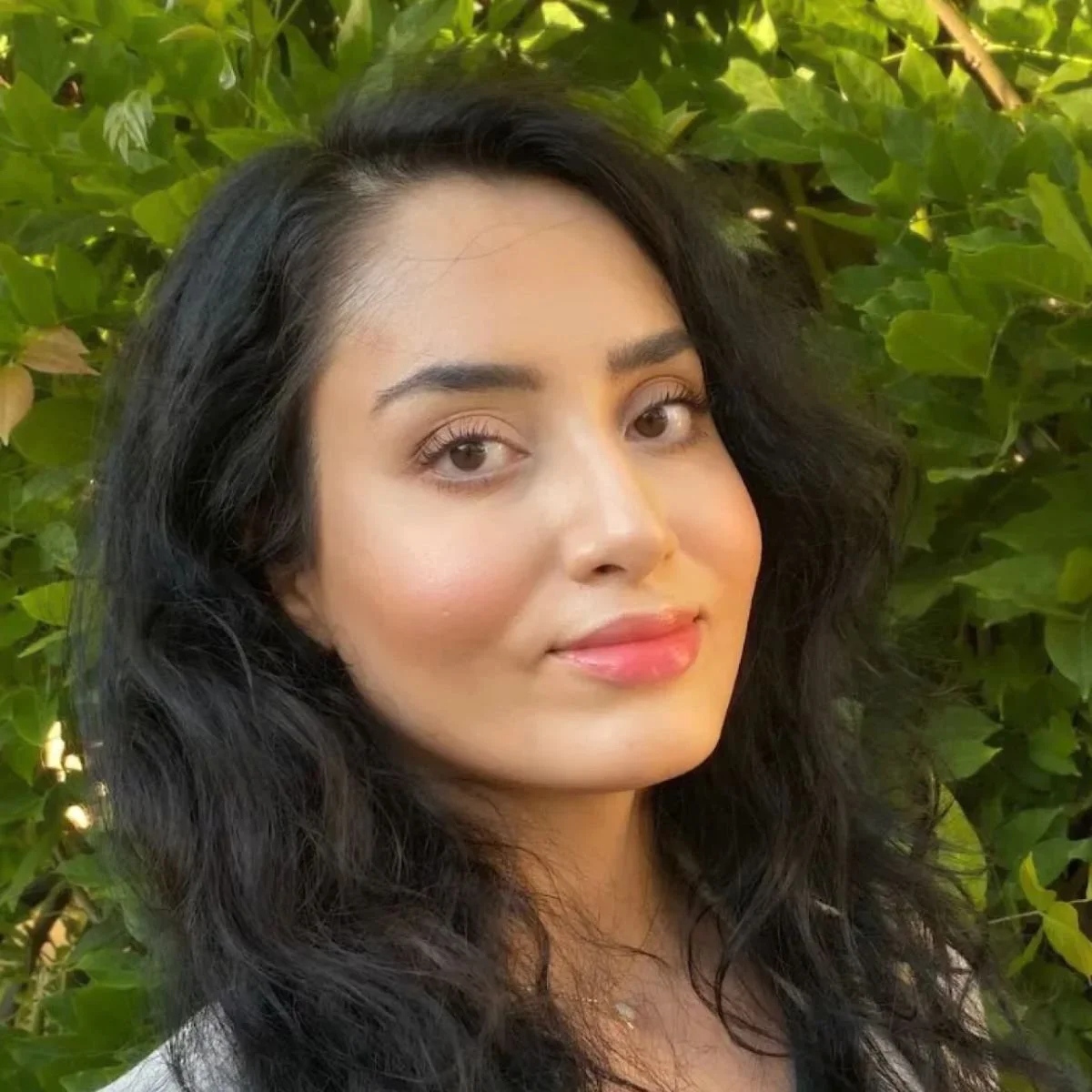 A woman with long black hair, light makeup, and a slight smile, standing outdoors with green leaves in the background.