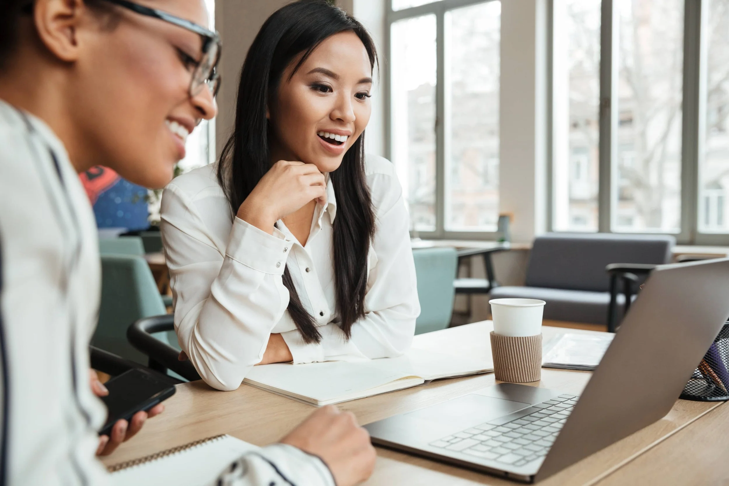 Two women sitting at a table in a bright office space, engaged in conversation, with laptops, a notebook, and a coffee cup on the table.