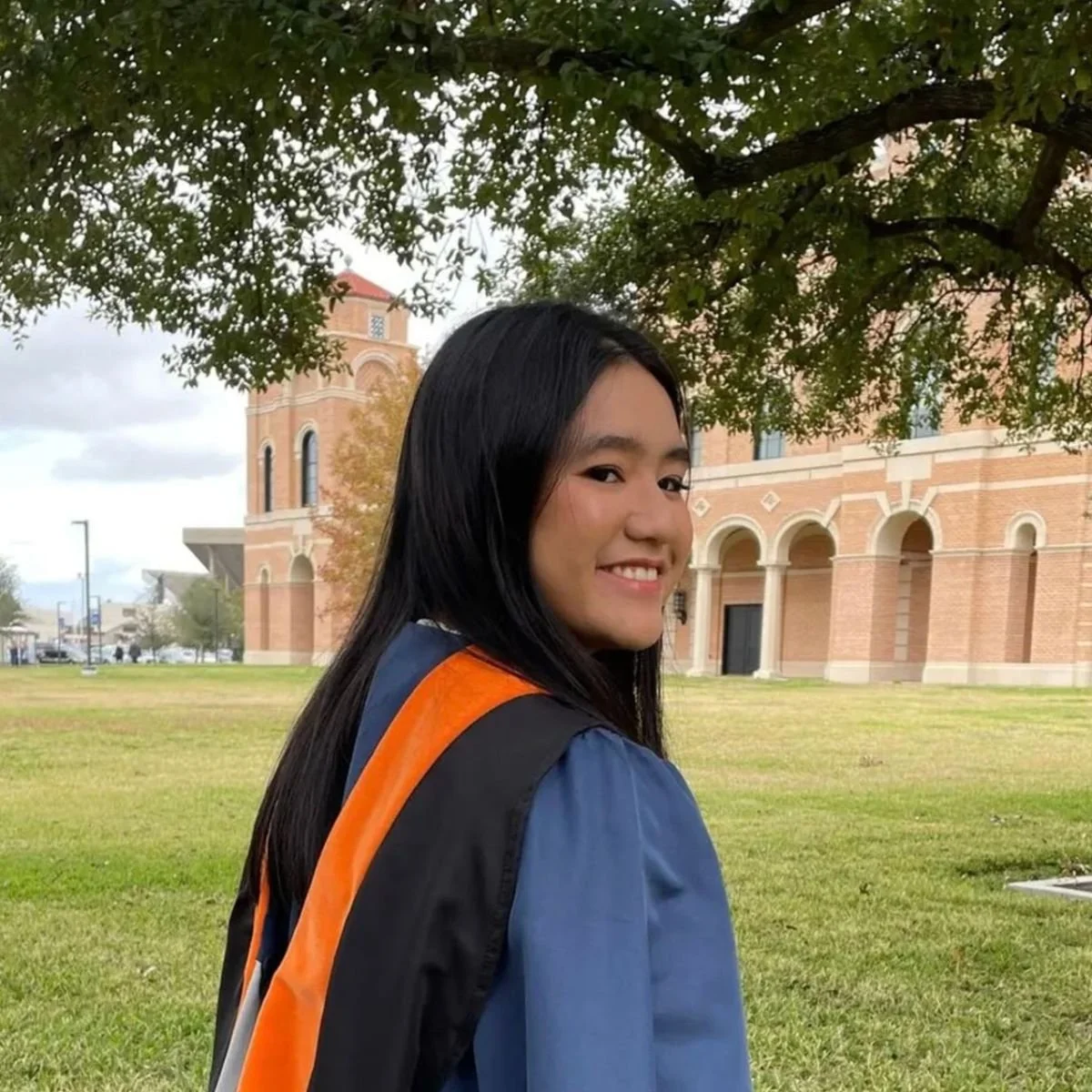 A young woman with long black hair smiling in a graduation gown and cap, standing on a grassy area with a large brick building and trees in the background.