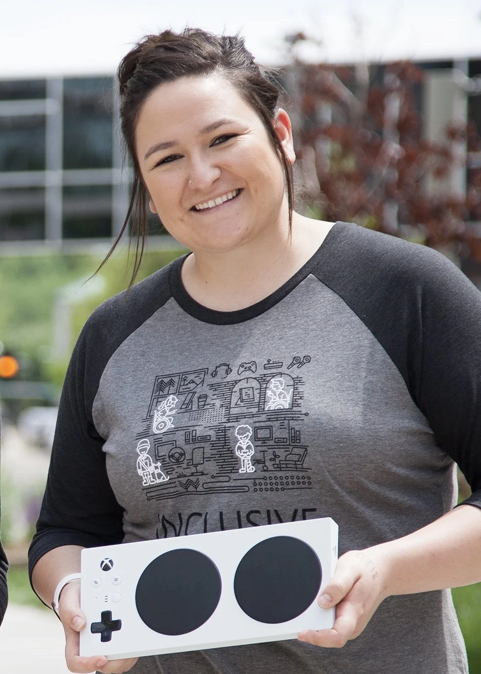 A caucasian woman with brown hair in an up-do is standing alone with a dark gray shirt on. She is holding a white xbox adaptive controller and is smiling.